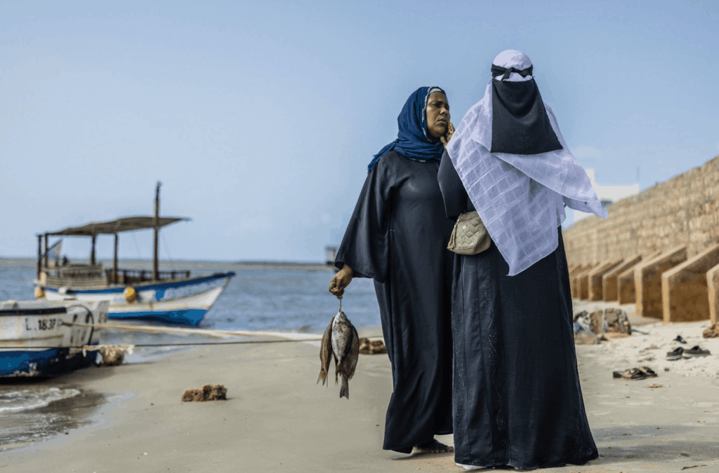 Women carrying fish on a beach with boats in the background